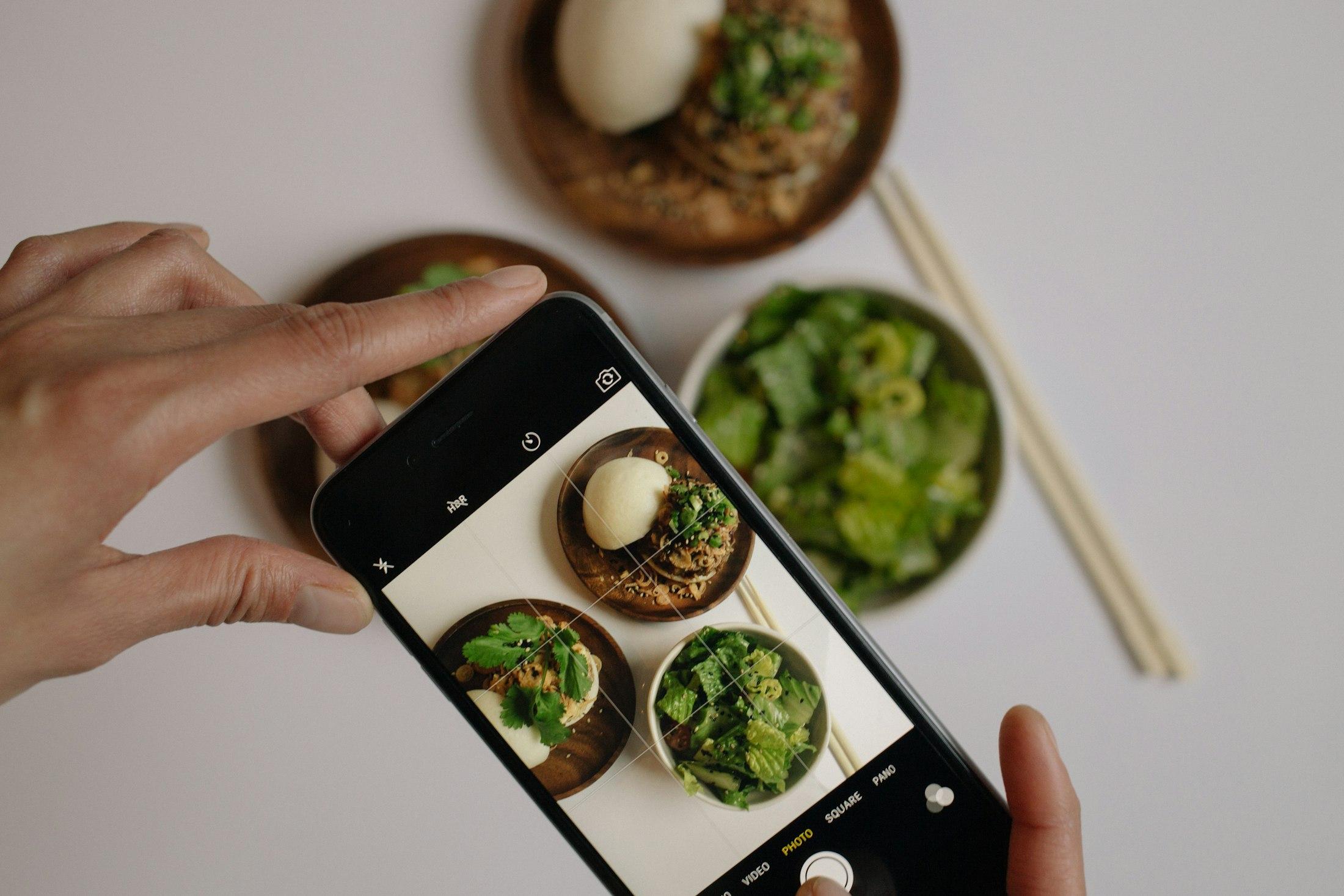 Someone photographing a bowl of noodles and greens with their phone